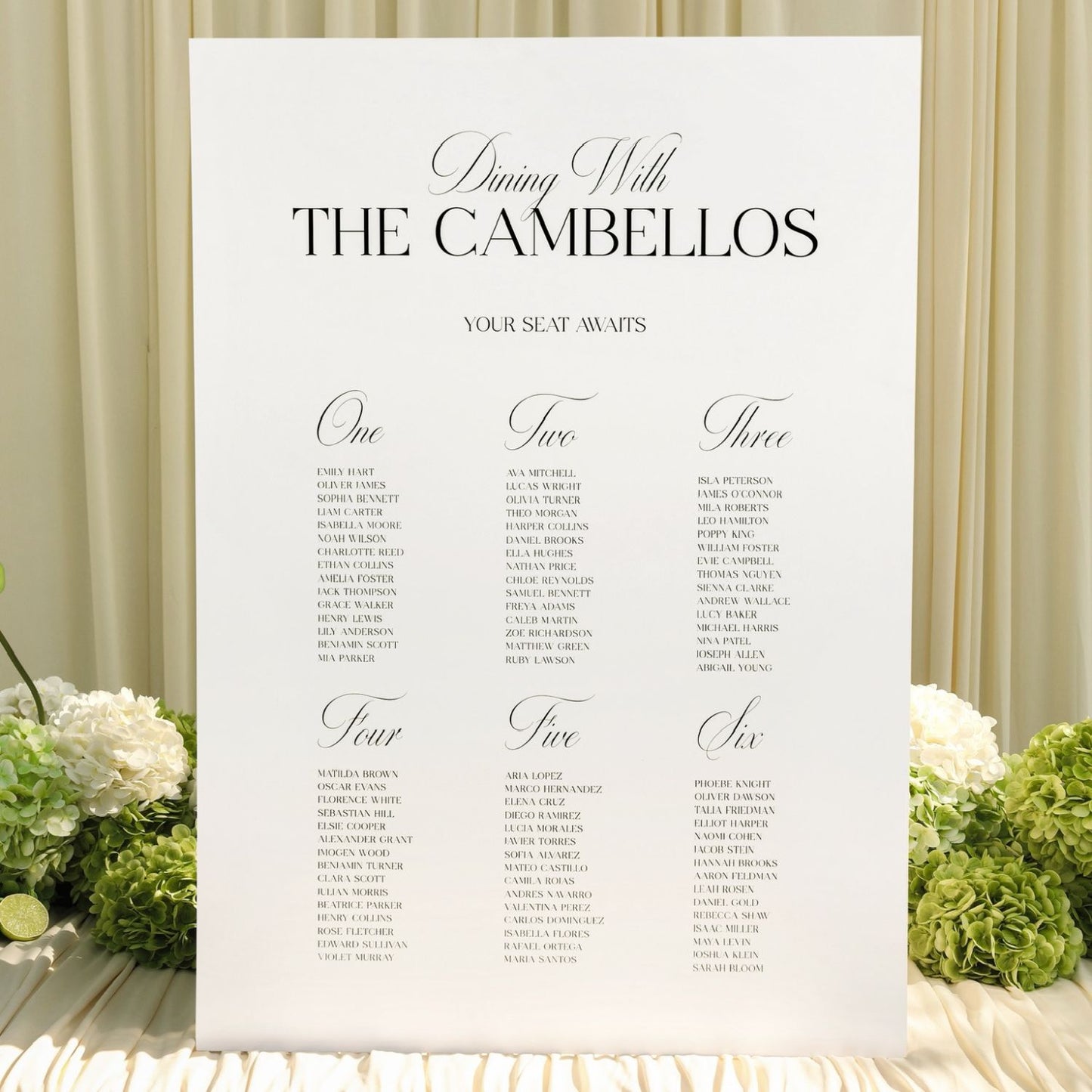A vertical white wedding seating chart sign reading 'Dining With THE CAMBELLOS' and listing seat numbers for guests, styled with white flowers and green hydrangeas against a cream draped backdrop.