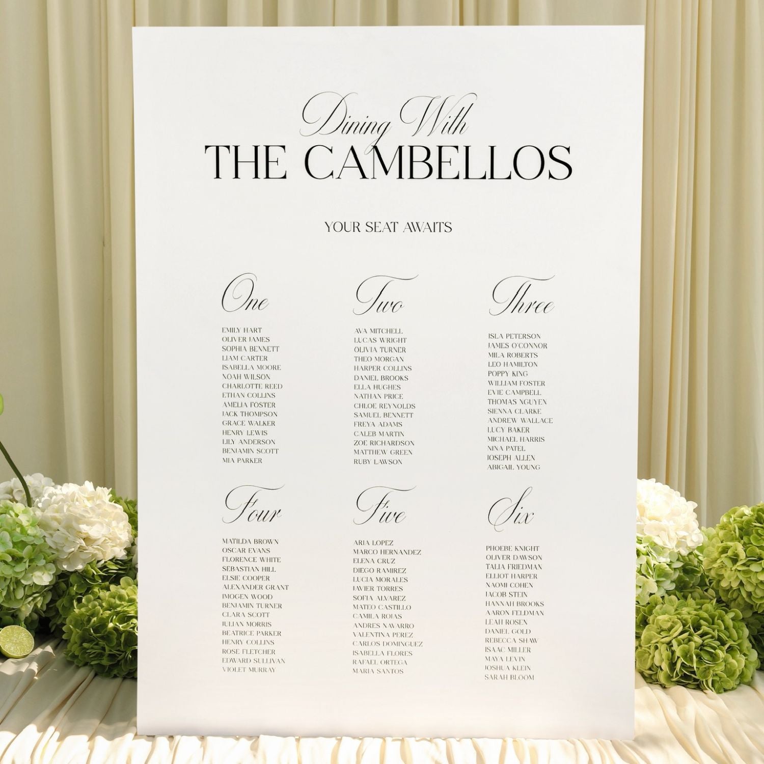 A vertical white wedding seating chart sign reading 'Dining With THE CAMBELLOS' and listing seat numbers for guests, styled with white flowers and green hydrangeas against a cream draped backdrop.